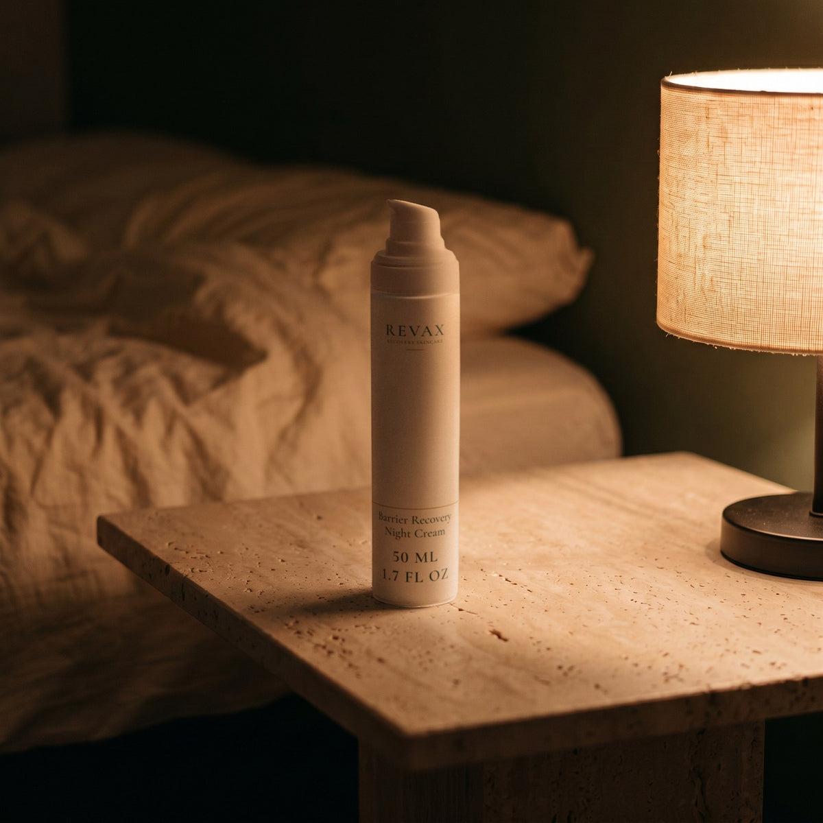 REVAX Barrier Recovery Night Cream 50 ml, pump bottle on a travertine nightstand beside a lit lamp at night, front view